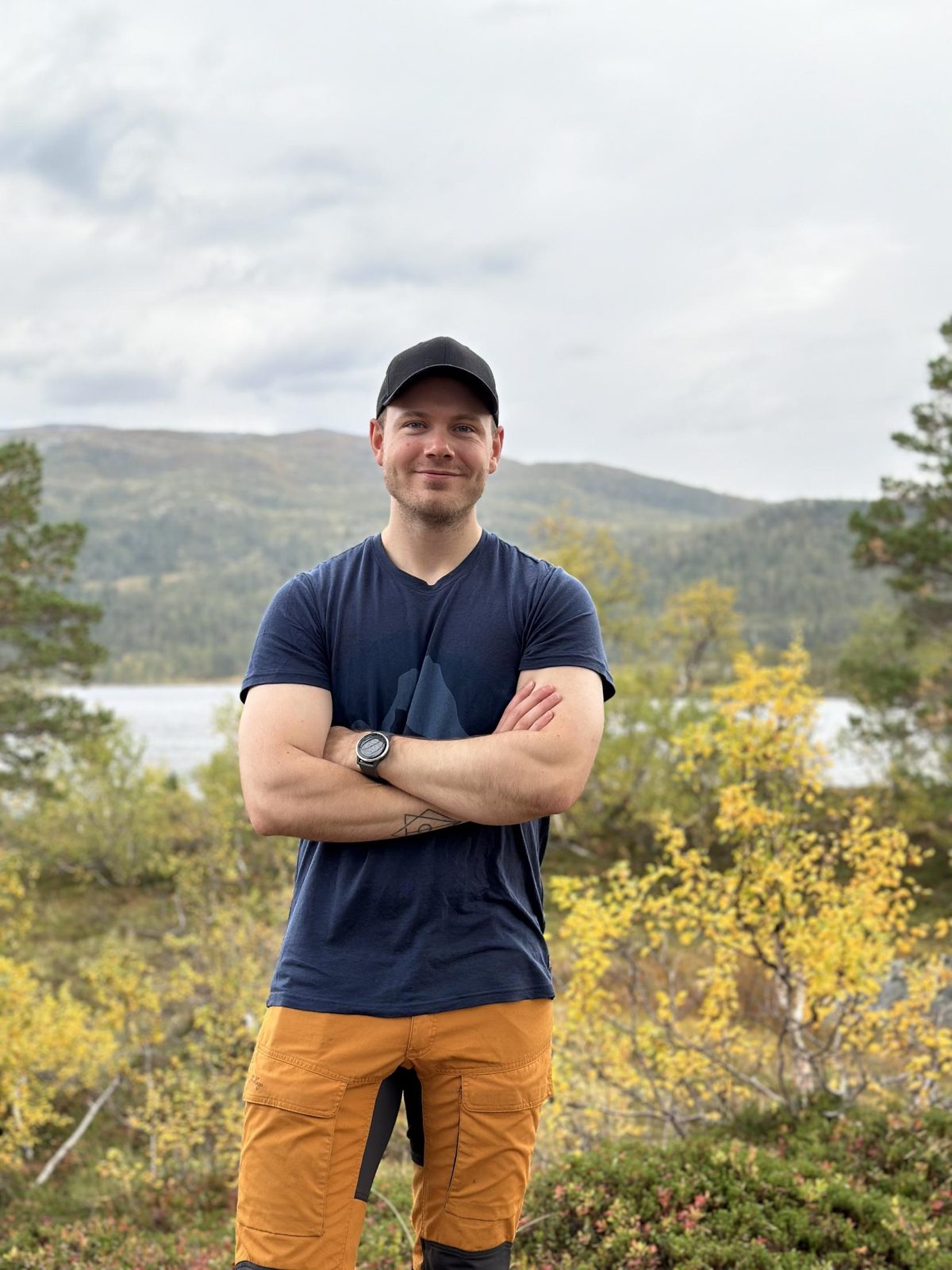 Marcus Larsen står med armene i kors foran et vann og fjell i høstfarger.